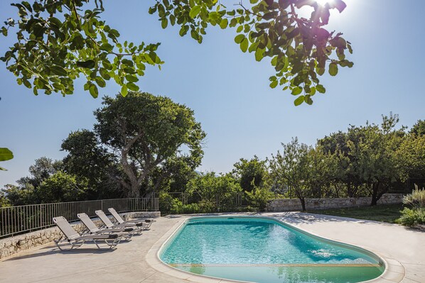 A swimming pool with lounge chairs, surrounded by trees and a stone wall.