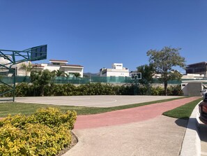 Uma quadra de basquete com cerca verde, palmeiras e edifícios ao fundo.