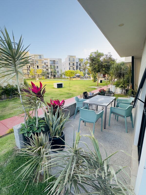 Uma varanda com mobiliário de exterior, plantas em vasos e vista para uma área residencial com edifícios e um jardim.