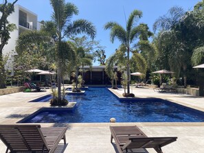 Uma área de piscina com espreguiçadeiras e guarda-sóis, rodeada por palmeiras e um céu azul límpido.