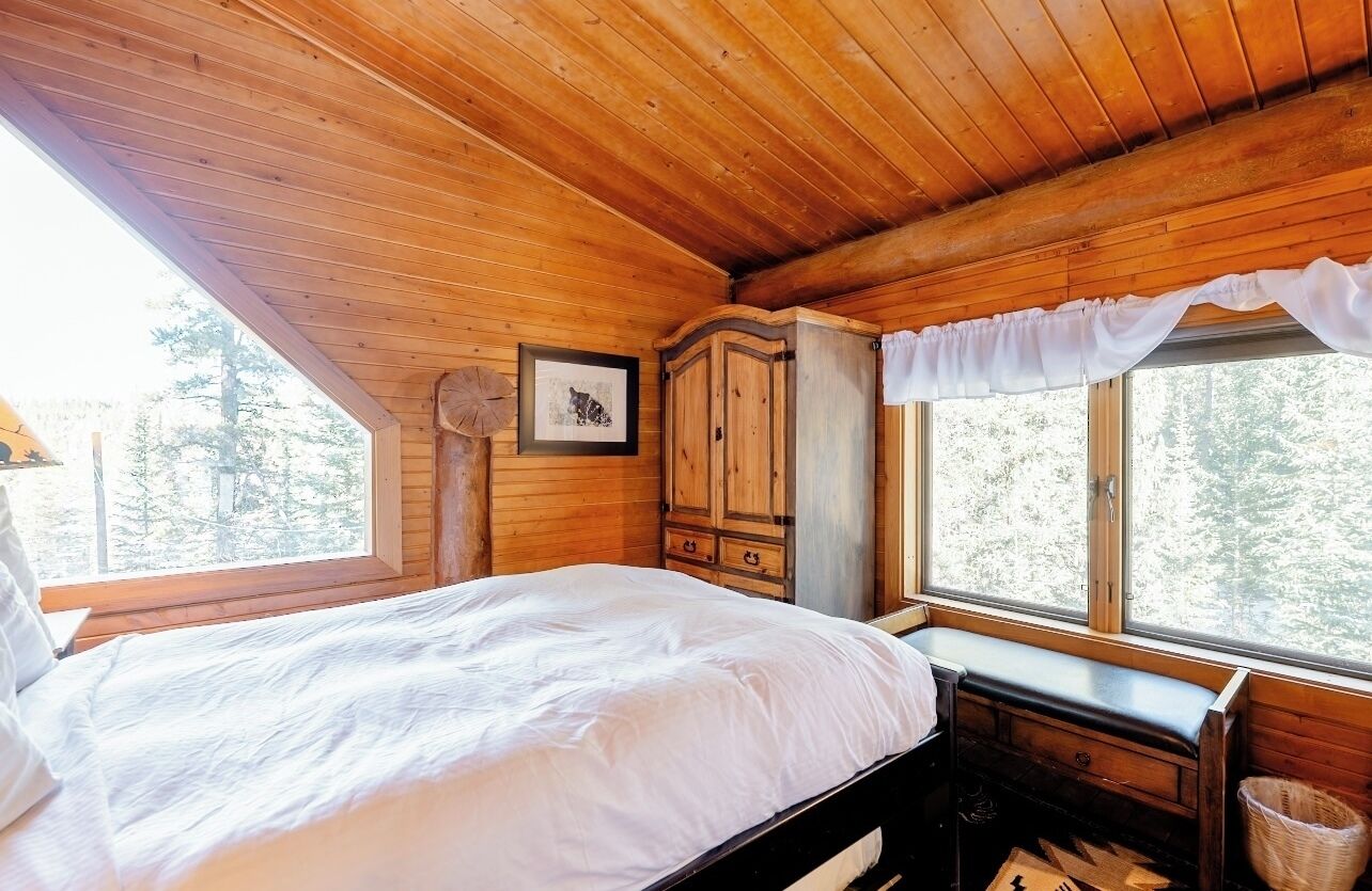 Ein Schlafzimmer einer Holzhütte mit Bett, Schrank, Fenster mit Vorhängen und Blick auf Bäume.