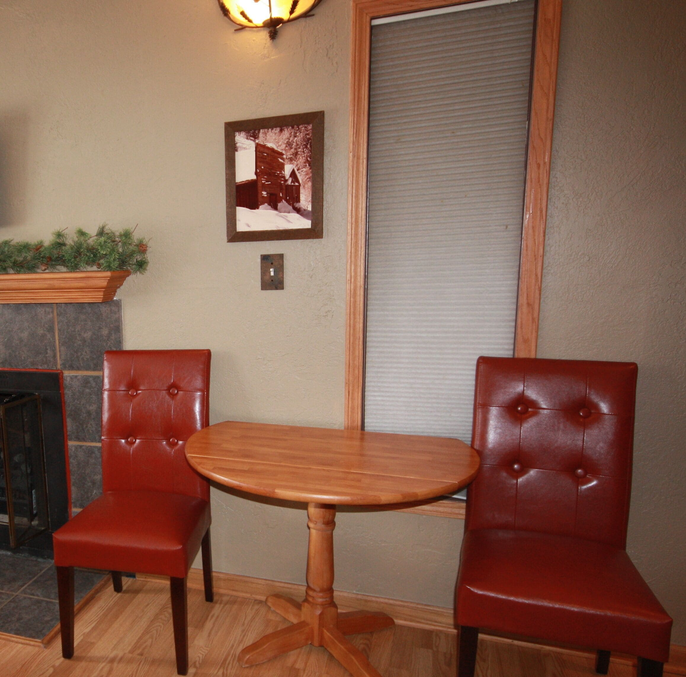 A room with two red leather chairs, a wooden table, a fireplace with a mantle, and a framed picture on the wall.