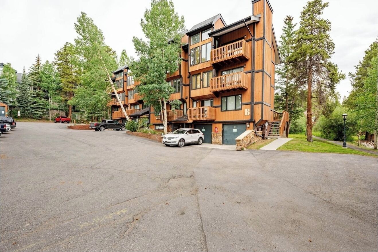 A multi-story wooden building with balconies, a parking area, and surrounding trees.