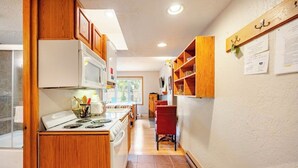 A compact kitchen with wooden cabinets, a microwave, and a stove. There is a dining area with a red chair and a wooden shelf with papers on the wall. - Four O'clock Lodge B05 (Breckenridge)
