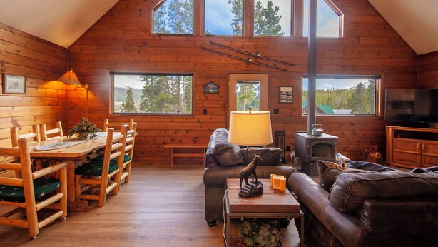 A cozy cabin interior with a wooden dining table, benches, a sofa, a fireplace, and large windows offering a view of the forest. - Haus Crisler (Breckenridge)