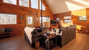 A log cabin interior with a loft bedroom, wooden walls, large windows, a sofa, a television, and a lamp. - Haus Crisler (Breckenridge)