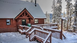 A wooden cabin with a snow-covered roof, a wooden porch, and a fenced deck. - Haus Crisler (Breckenridge)