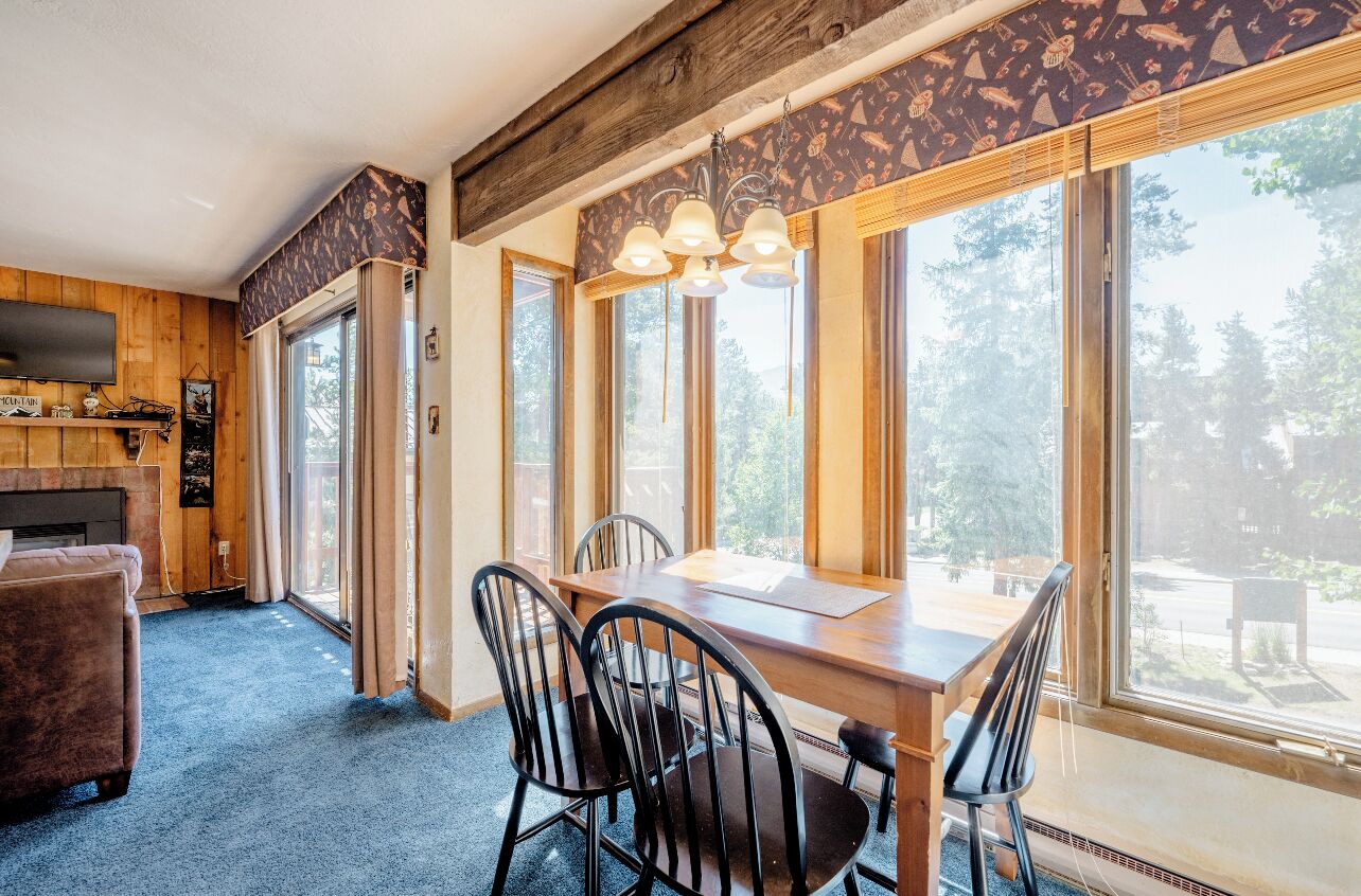 A cozy room with a wooden dining table, four chairs, and a television on the left. Large windows provide a view of the outdoors.
