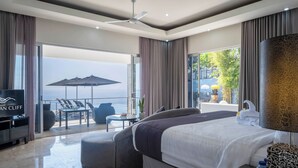 Una habitación de hotel moderna con cama, televisión, balcón con vistas al mar y zona de comedor con mobiliario exterior.