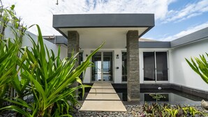 Un edificio moderno con una galería techada que conduce a una puerta de cristal, rodeado de plantas diversas y un pequeño elemento acuático.