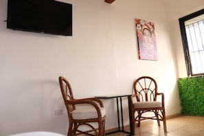 A room with two wicker chairs, a small table, a flat-screen TV mounted on the wall, and a window with a green wall.