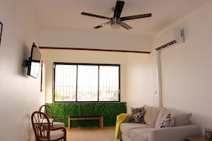 A living room with a sofa, a wooden bench, a dining table with chairs, a ceiling fan, and a large window with a view of greenery outside.