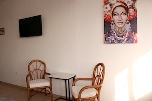 A room with two wicker chairs and a small table, a painting on the wall, and a television mounted on the wall.