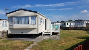 A mobile home with a covered porch and a small deck, surrounded by a grassy area and a wooden fence. - 8 Berth Caravan with Spacious enclosed garden (Brean)