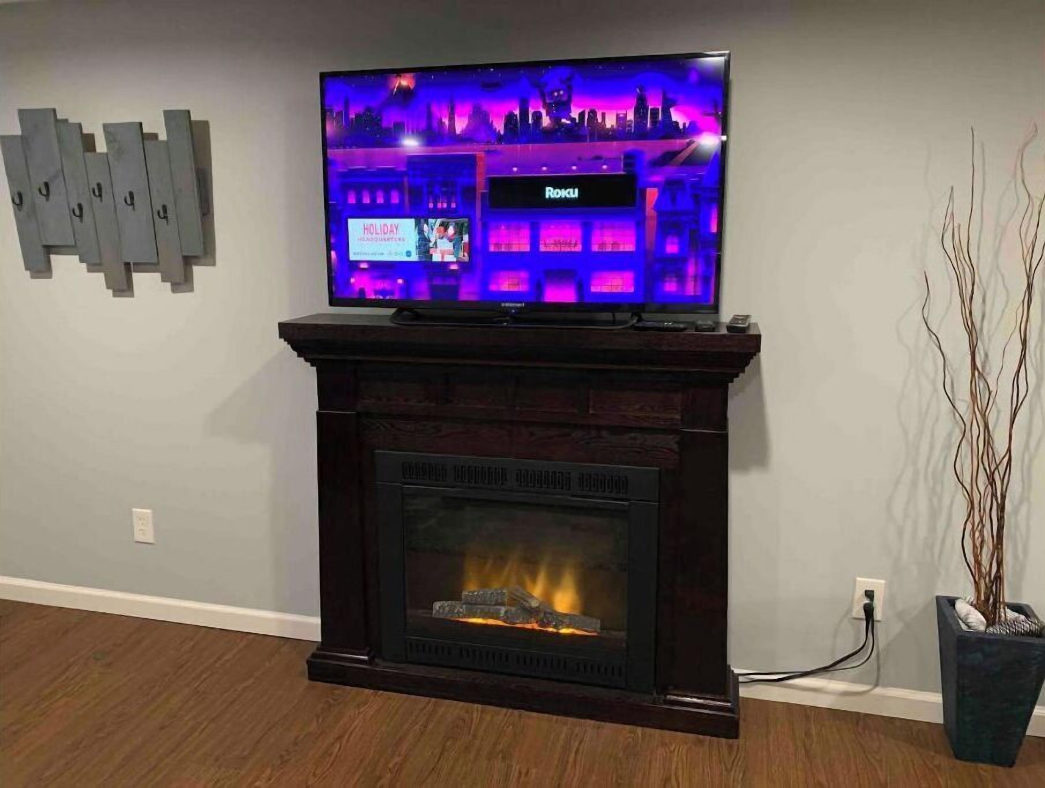 Un téléviseur à écran plat est posé sur une cheminée en bois, au-dessus d’un foyer où l’on voit des flammes.