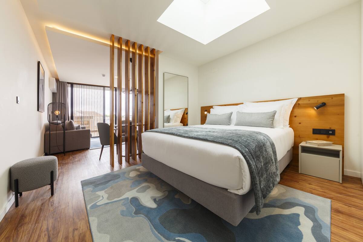 A modern hotel room with a large bed, a small bedside table, a chair, a sofa, a large window, and a skylight.