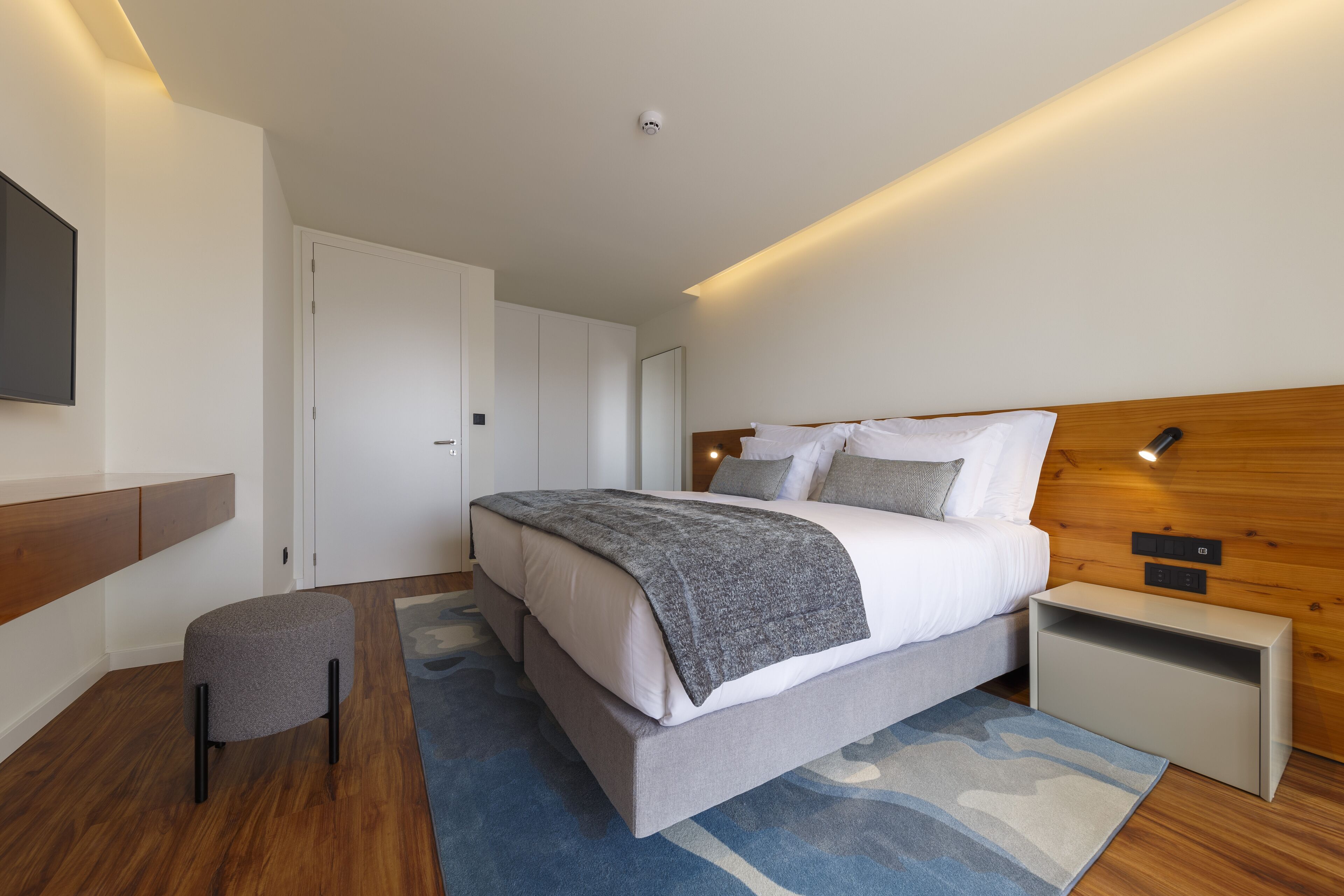 Ein modernes Schlafzimmer mit einem großen Bett, einem Nachttisch, einem kleinen Hocker und einem flachbildfernseher an der Wand.
