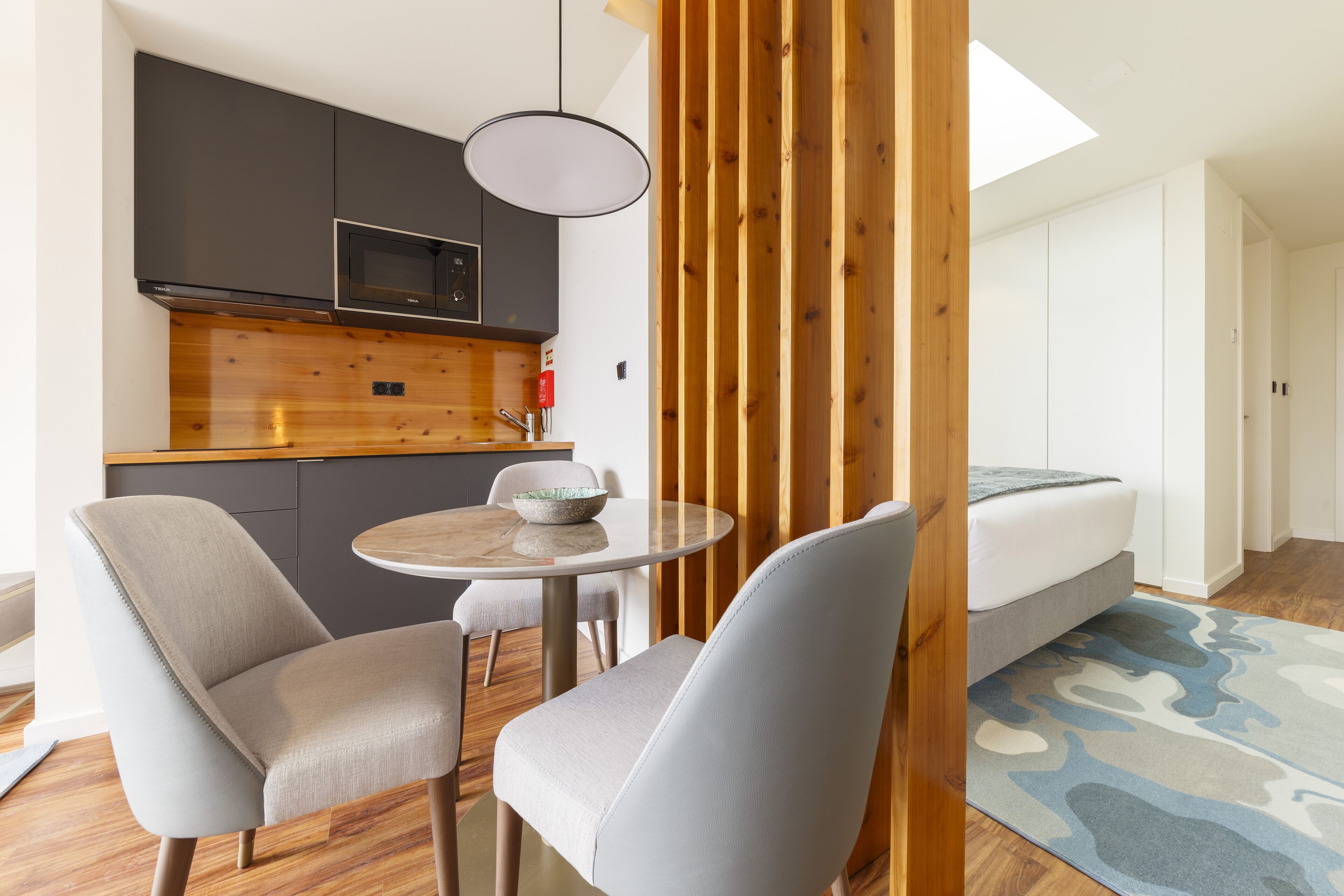 A compact kitchen with modern appliances, a dining area with two chairs, and a bed with a blue patterned rug.