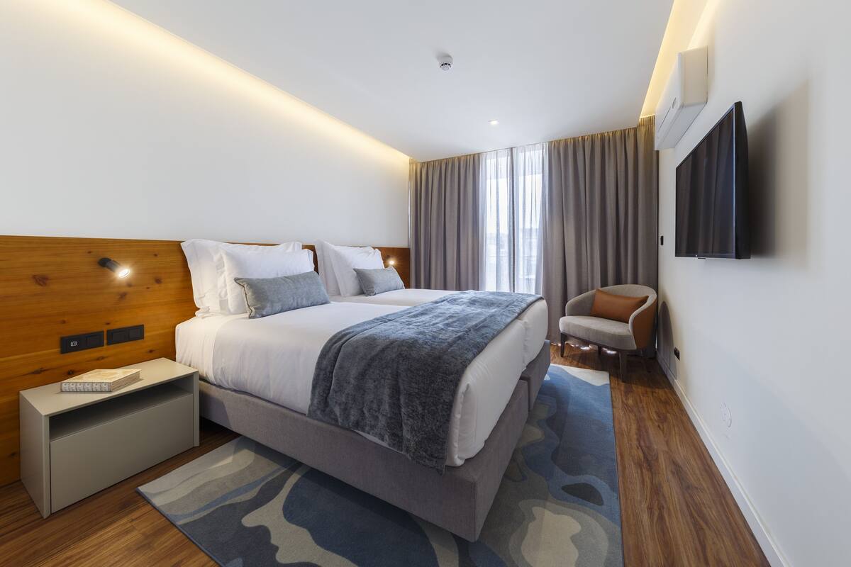 A modern hotel room with a large bed, a flat-screen TV mounted on the wall, a small side table, and a view of the city through large windows.