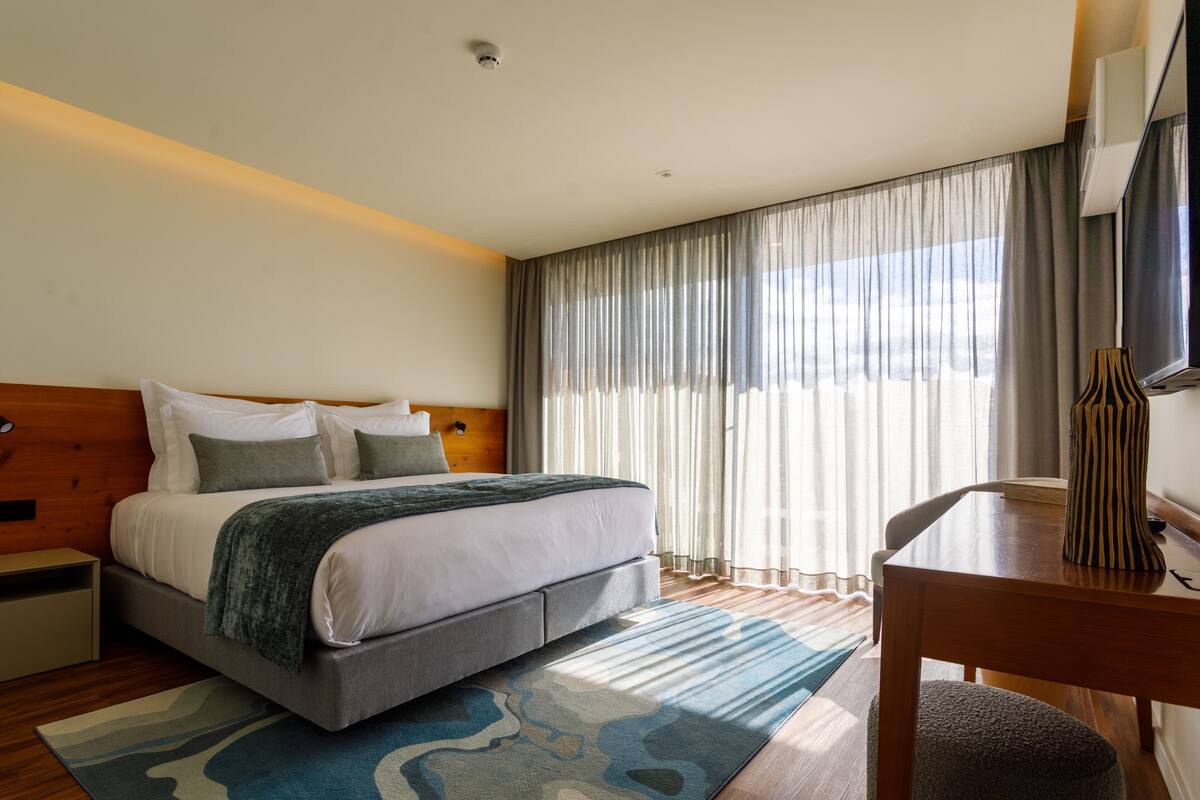 A modern hotel room with a large bed, a wooden headboard, a desk with a chair, and a large window with sheer curtains.