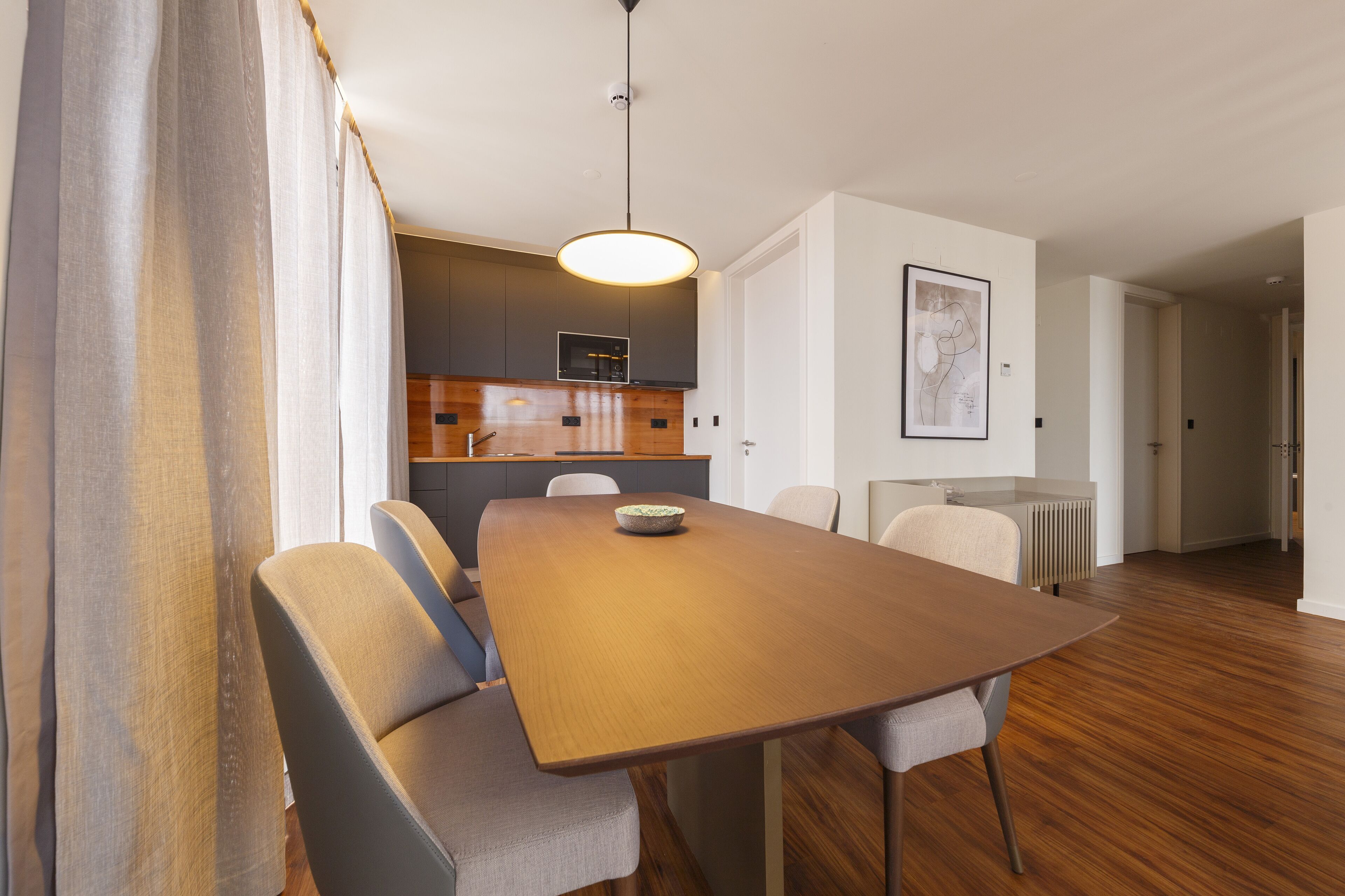 A modern dining area with a wooden table and cushioned chairs, a pendant light, and a kitchen in the background.