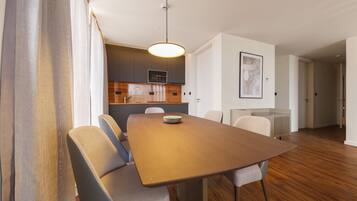 A modern dining area with a wooden table and cushioned chairs, a pendant light, and a kitchen in the background.