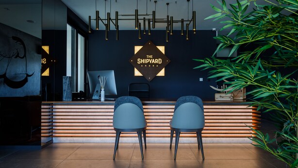 A reception area with a modern design, featuring a dark wall with a geometric sign, two grey chairs, a wooden reception desk, and a potted plant. - The Shipyard Angra (Angra do Heroismo)