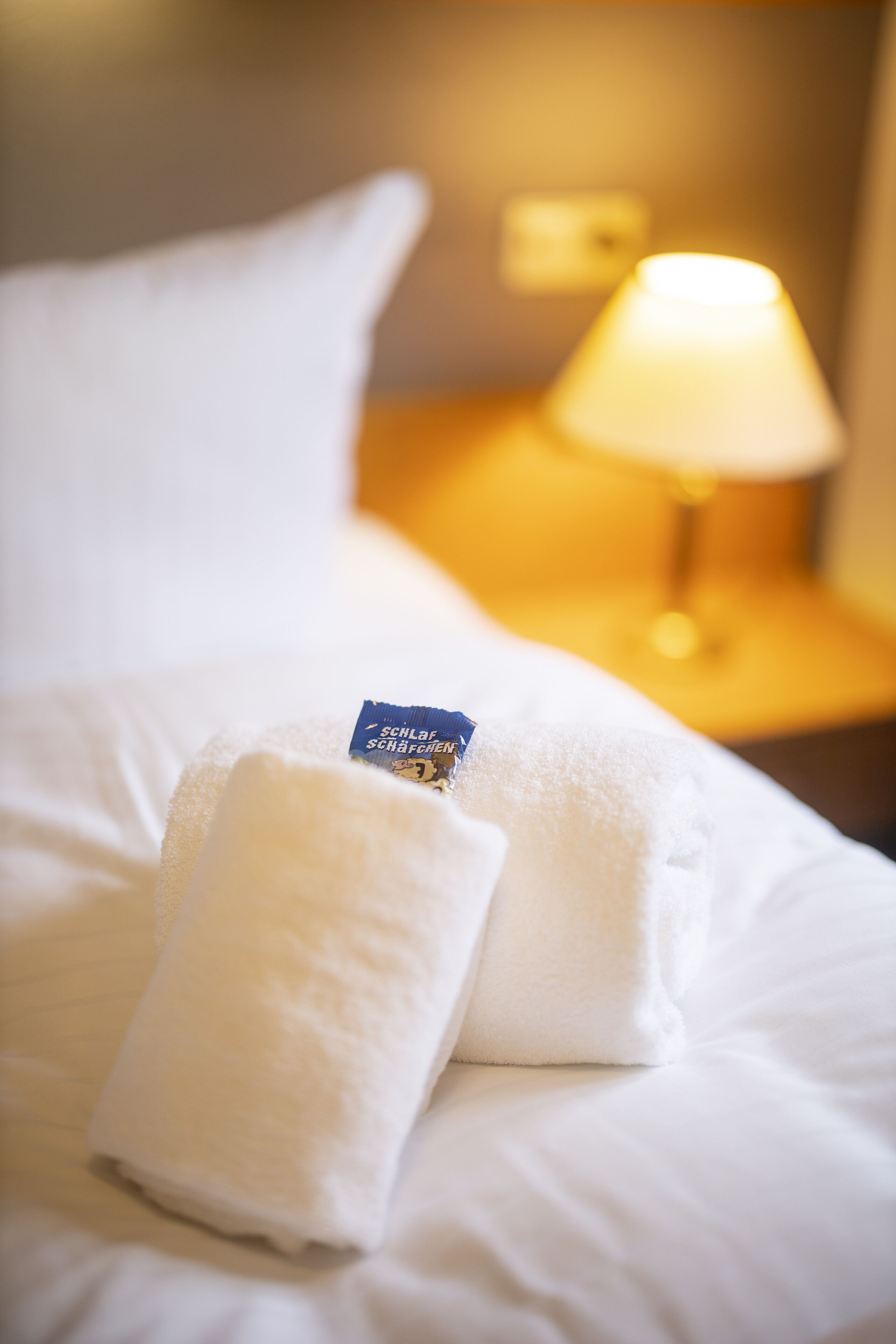 A neatly made bed with white linens and pillows, a small blue tag on one pillow, and a lamp on a bedside table in the background.