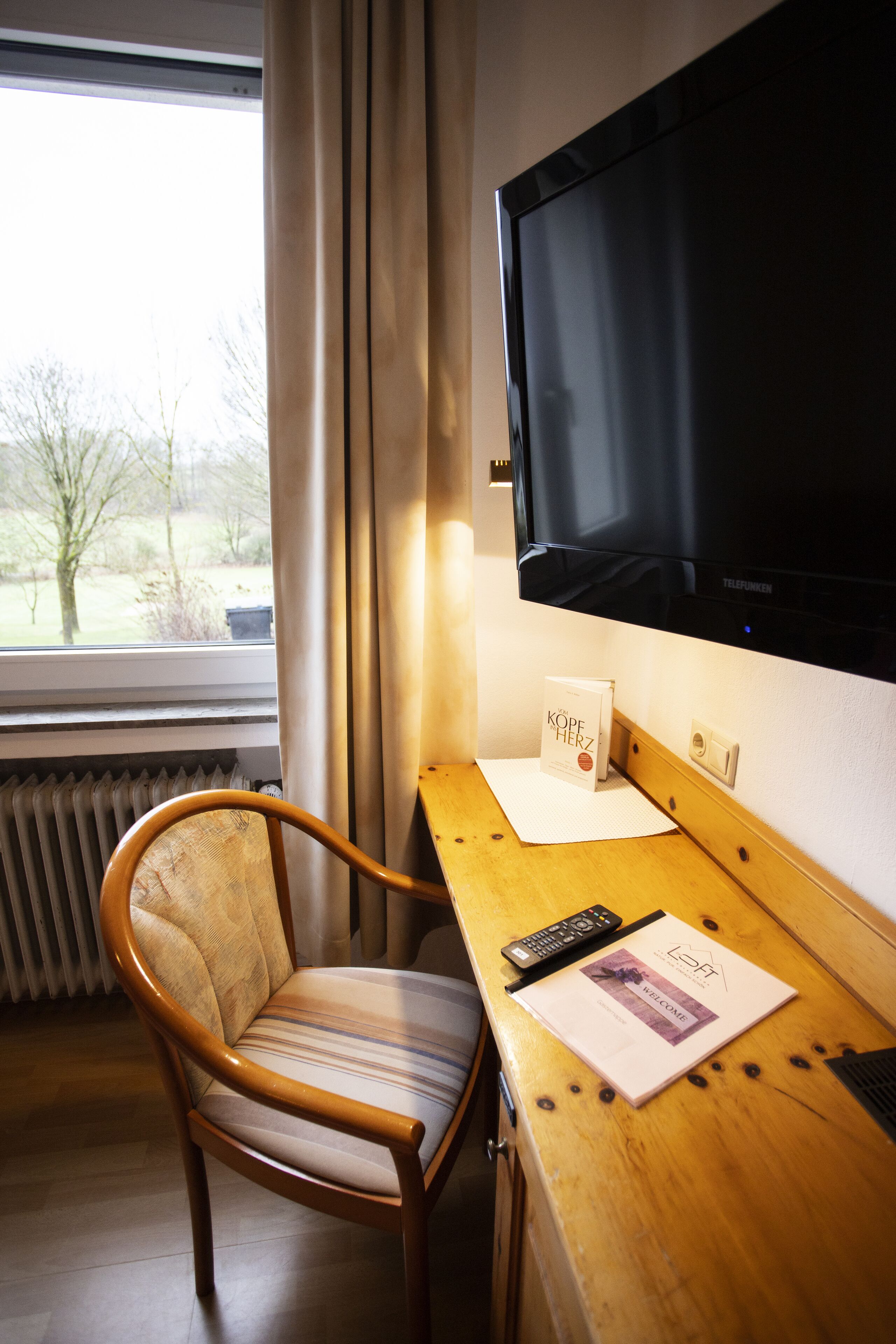A hotel room with a wooden desk, a chair, a flat-screen TV mounted on the wall, a radiator, and a window with curtains.