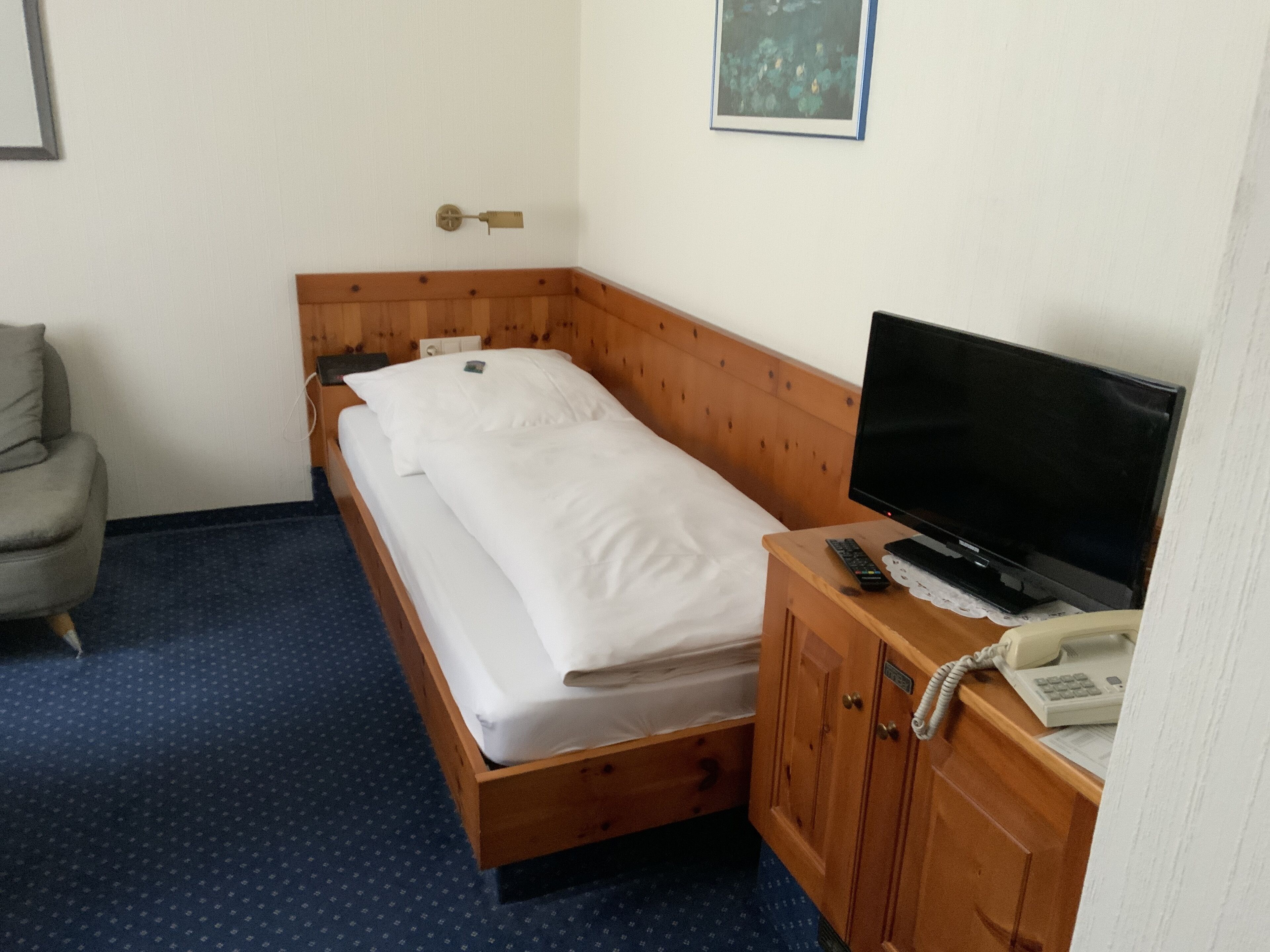 A single bed with white linens, a wooden headboard, a television on a wooden stand, and a telephone on the stand.
