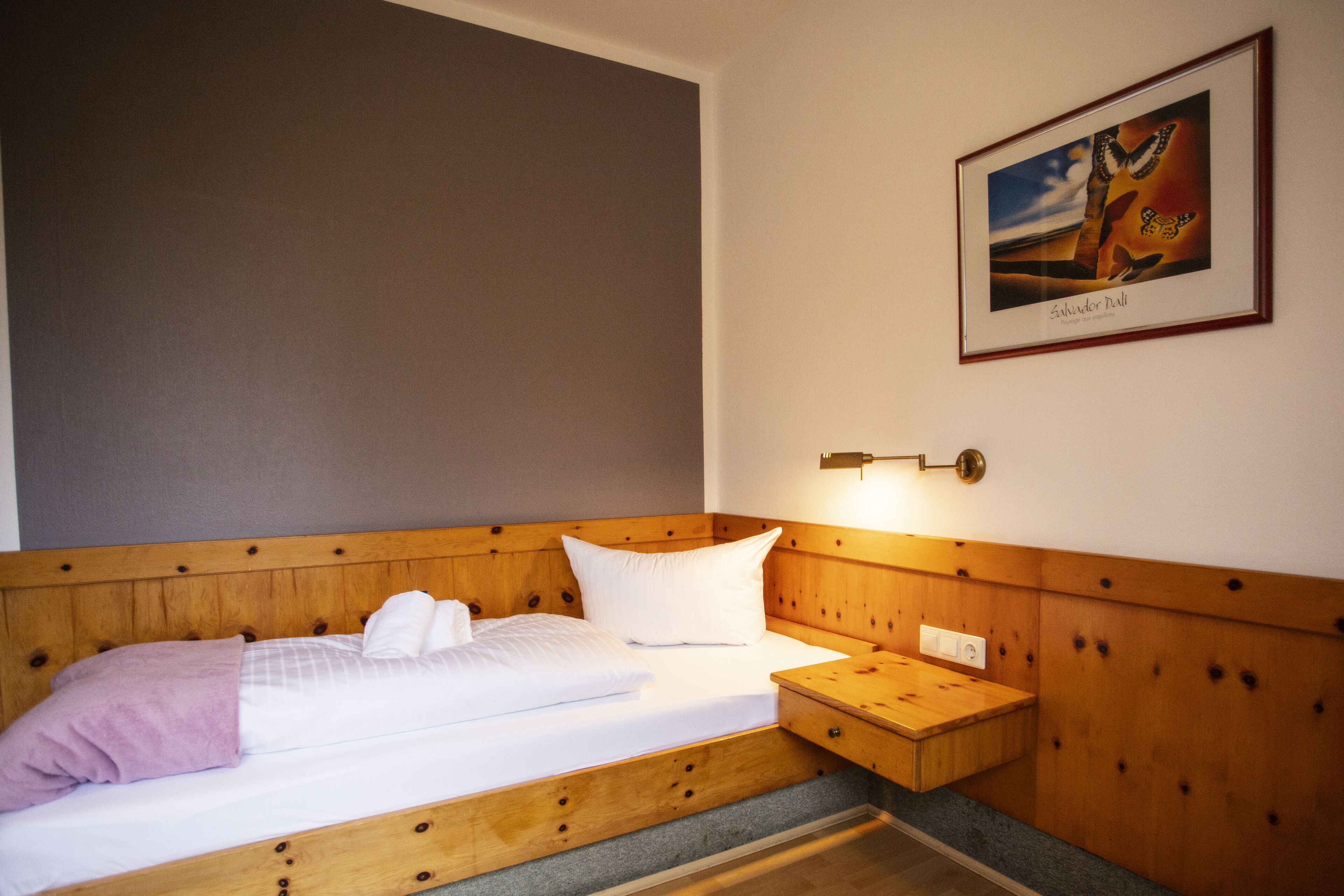 Cama bem arrumada com lençóis brancos e um cobertor roxo, cabeceira de madeira, mesa de cabeceira com abajur e um quadro com uma paisagem na parede.
