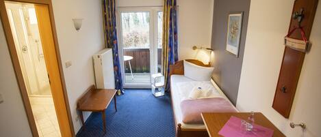 A small hotel room with a bed, a desk, a chair, a table, a radiator, a window with curtains, a door, and a wall-mounted shelf with a picture.