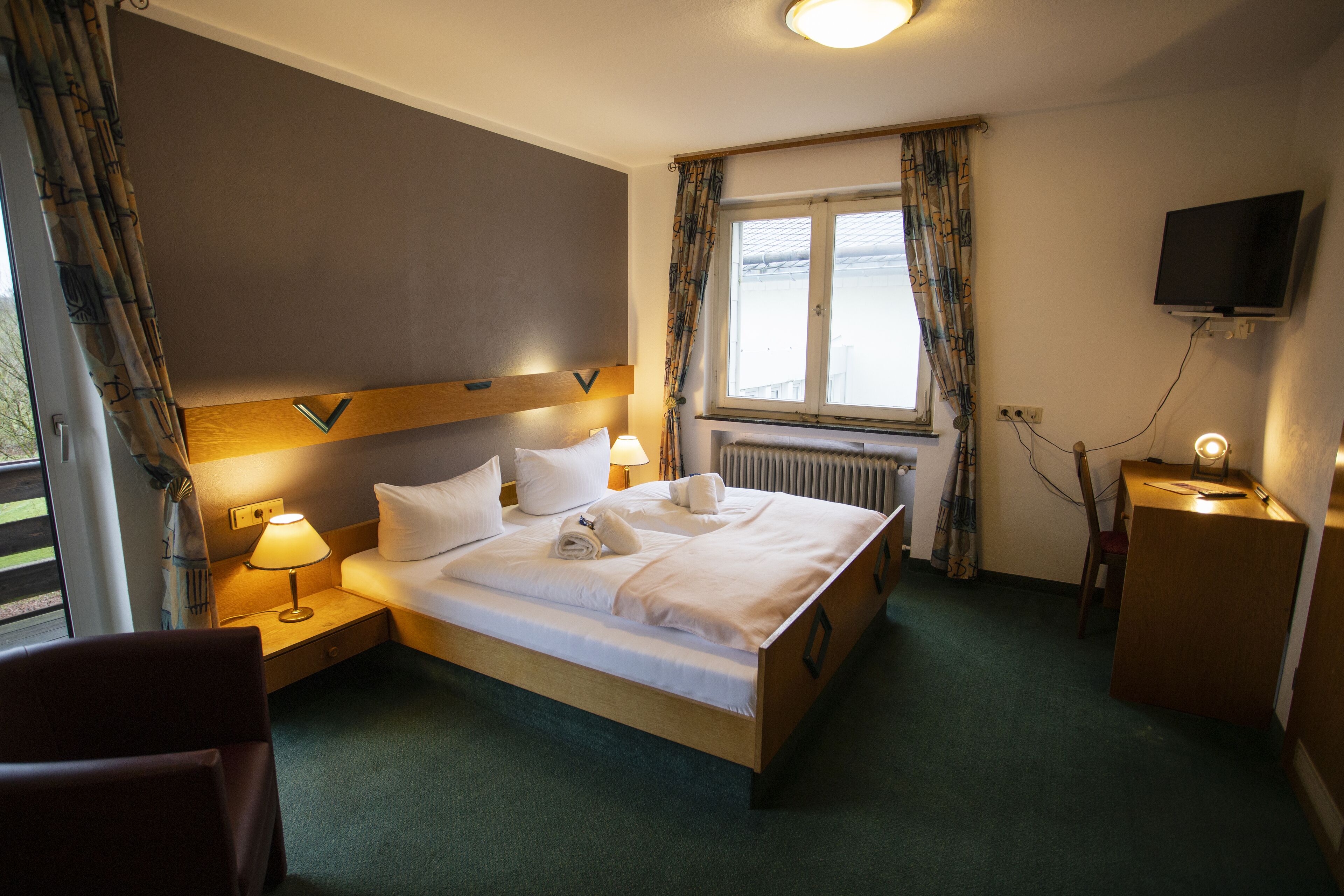 A hotel room with a bed, bedside lamps, a TV, and a window with curtains.