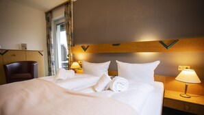 Quarto de hotel com uma cama grande, duas mesas de cabeceira com luminárias, uma cabeceira de madeira, uma cadeira e uma janela com cortinas.