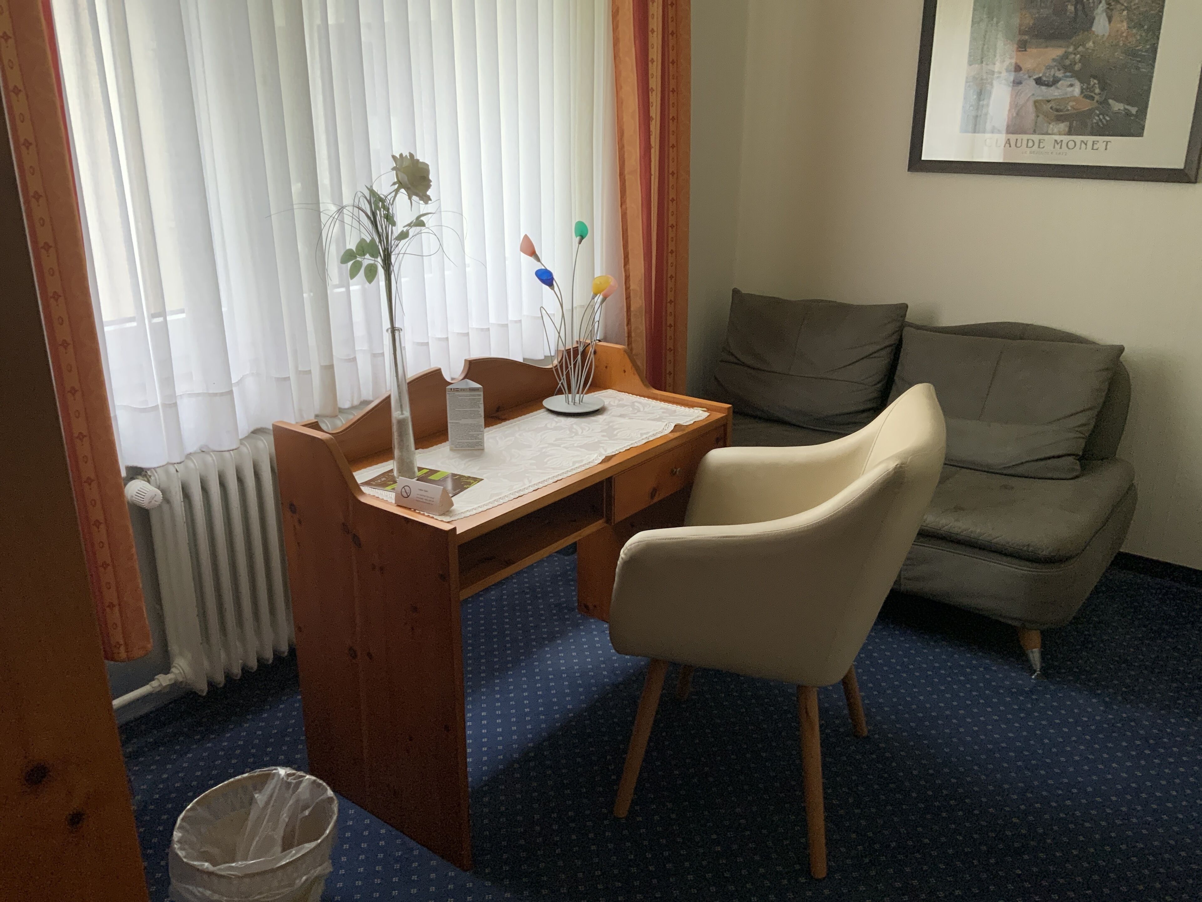 A room with a wooden desk, a chair, a radiator, a framed picture on the wall, and a grey sofa.