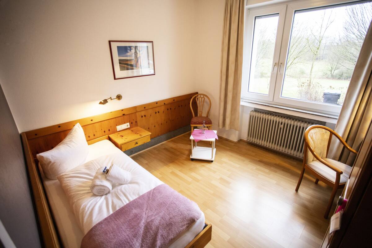 A bedroom with a wooden bed, a chair, a small table, and a window with a view of trees.