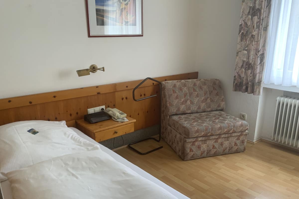 A hotel room with a single bed, a bedside table with a telephone and a book, a chair, and a radiator.