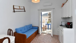 A compact living space with a blue sofa, a dining table, a kitchen area with a refrigerator, and a view through an open door to the outside. - Kastro Traditional House (Paros)