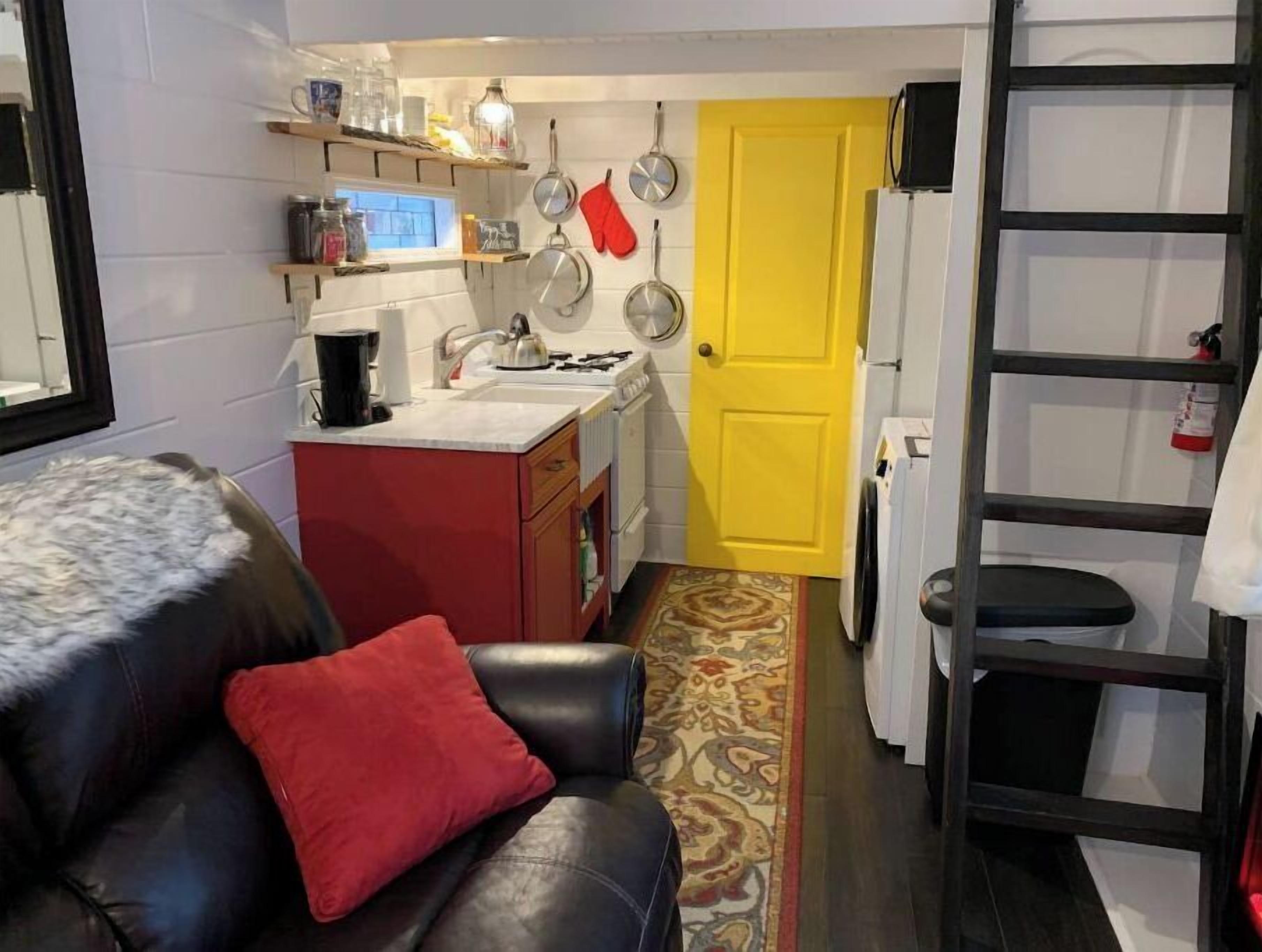 Un espace de vie compact comprenant un coin cuisine, un canapé avec un coussin rouge, un tapis à motifs et une porte jaune.