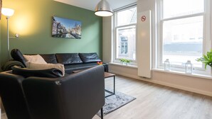 Ein modernes Wohnzimmer mit einer schwarzen Ledersofa, einer Stehlampe, einem Stadtbildgemälde, einem großen Fenster mit Blick und einem kleinen Tisch mit Pflanze.