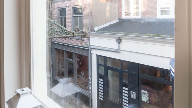 Blick aus dem Fenster auf ein Gebäude mit Backsteinfassade, ein Geschäft mit Schaufenster und ein Metallgeländer im oberen Stockwerk.
