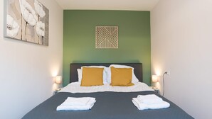 Ein ordentlich bezogenes Bett mit weißen und senffarbenen Kissen, einer dunkelblauen Bettdecke und zwei gefalteten Handtüchern.