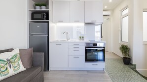 Una cocina moderna con armarios blancos, microondas integrado, refrigerador y horno. Hay un sofá gris con un cojín decorativo y una planta en maceta sobre la encimera.