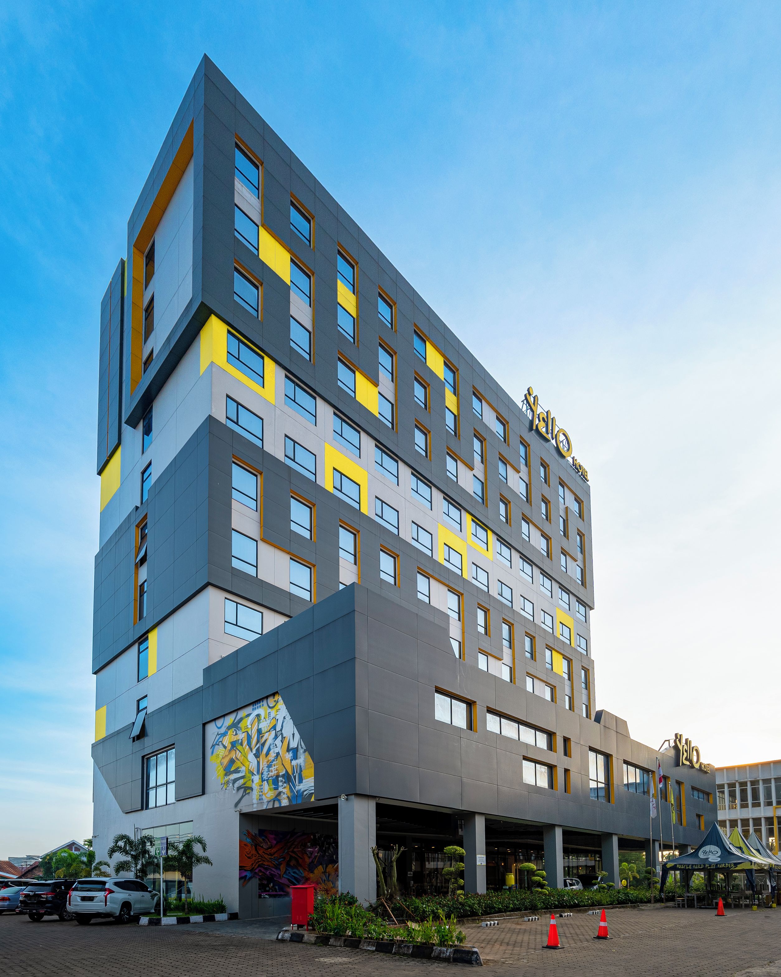 Un hôtel à plusieurs étages doté d’une architecture moderne, avec une entrée imposante ornée d’une fresque colorée, un espace de stationnement et un ciel dégagé.