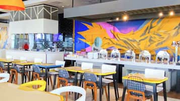 A modern cafe interior with multiple tables and chairs, a counter with a beverage display, and a colorful mural on the wall.