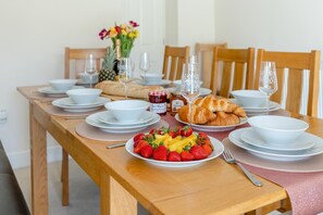 A dining table set with plates, bowls, a fruit platter, croissants, and wine glasses. - 4 Bed in Mortehoe (oc-dillyd) (Mortehoe)