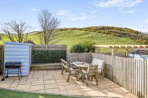 A patio with a grill, wooden furniture, and a view of a hilly landscape. - 4 Bed in Mortehoe (oc-dillyd) (Mortehoe)