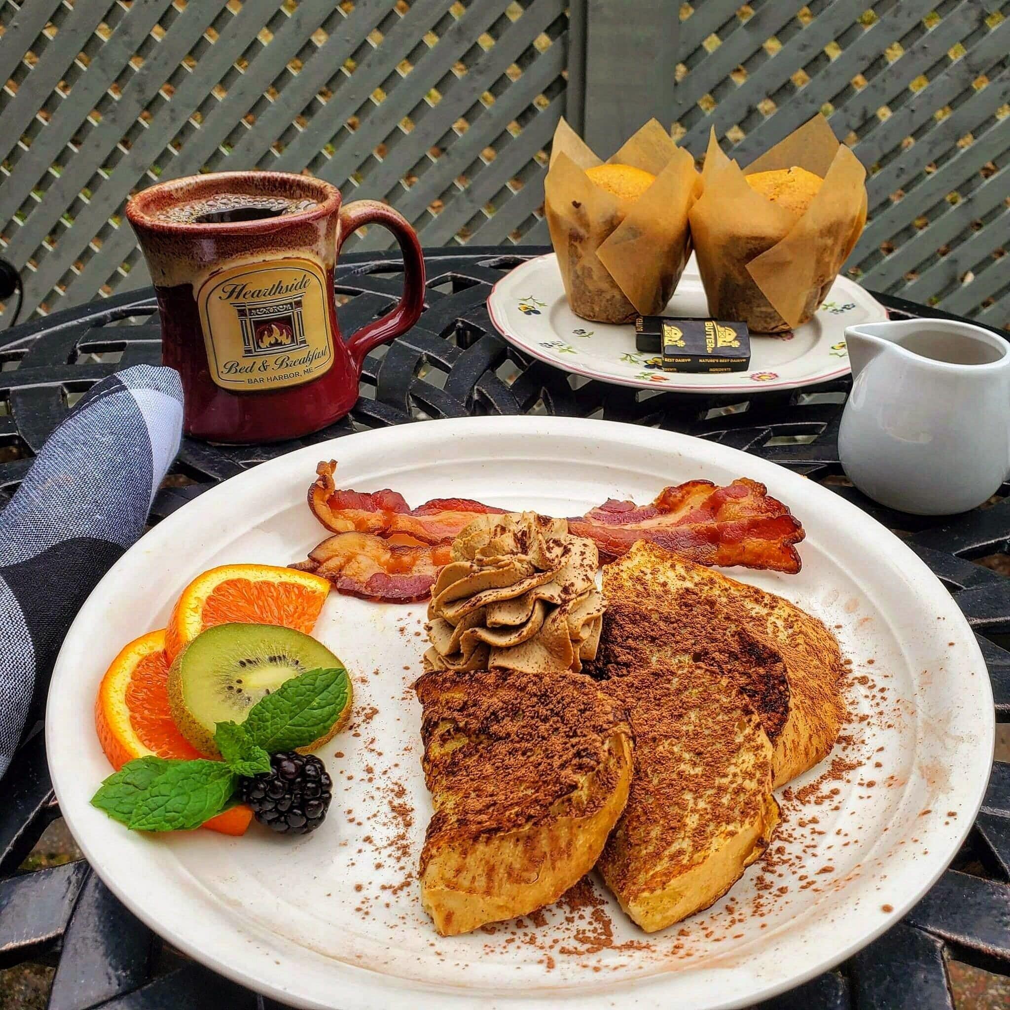 Une assiette de petit-déjeuner avec des œufs, du bacon, des champignons et des fruits, posée sur une table avec une tasse de café et une assiette de viennoiseries.