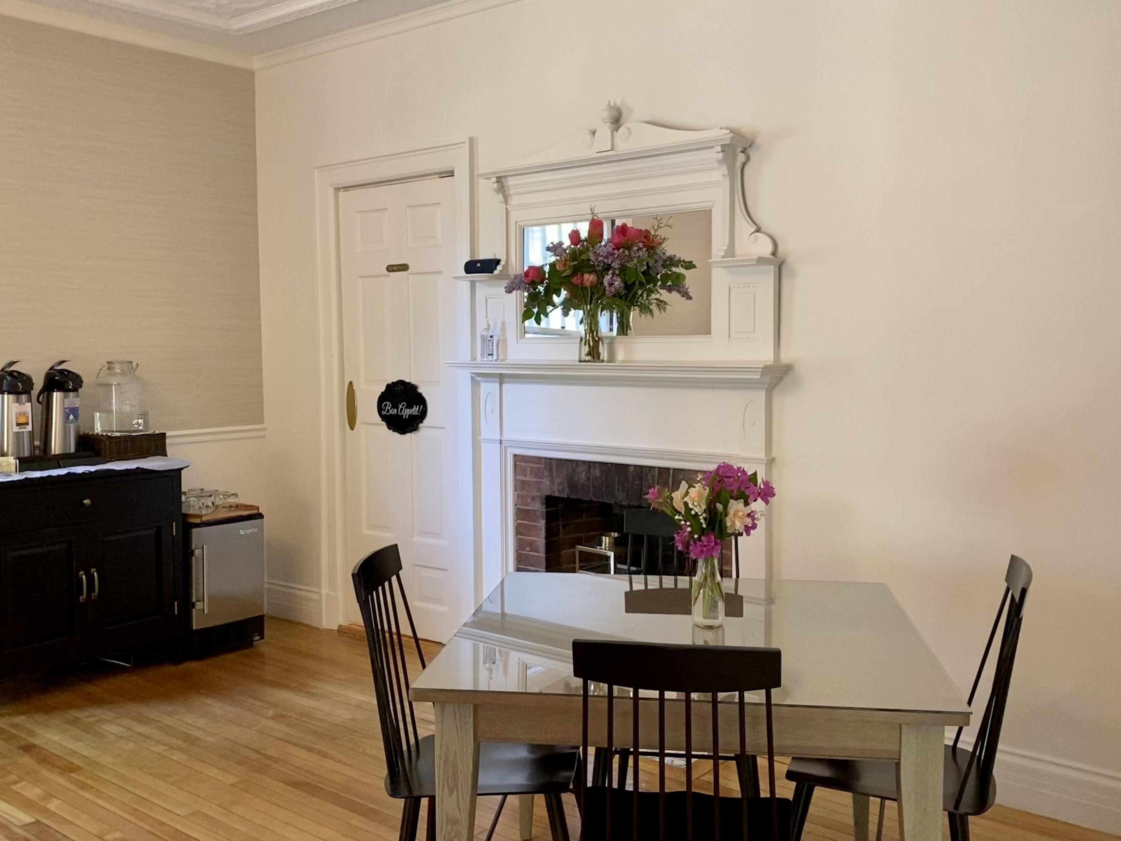 A dining area with a white fireplace, a table, and chairs. There is a door with a sign that reads "Best Appetites!" and a refrigerator in the background.