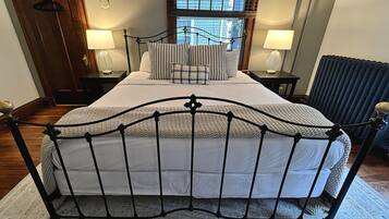 A neatly arranged bedroom with a metal bed frame, a large window with blinds, two bedside tables with lamps, and a radiator.